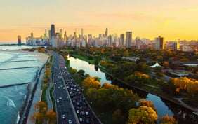 Scenic aerial view of the Chicago skyline at sunset, with busy Lake Shore Drive running alongside Lake Michigan and autumn trees framing the distant skyscrapers, including the John Hancock Center. - Saviynt
