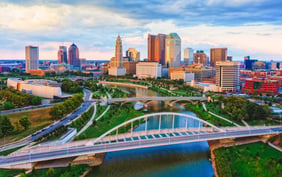 Vibrant aerial view of the Columbus skyline featuring the Art Deco LeVeque Tower and downtown skyscrapers rising above the Scioto River and the arched Main Street Bridge at golden hour. - Saviynt