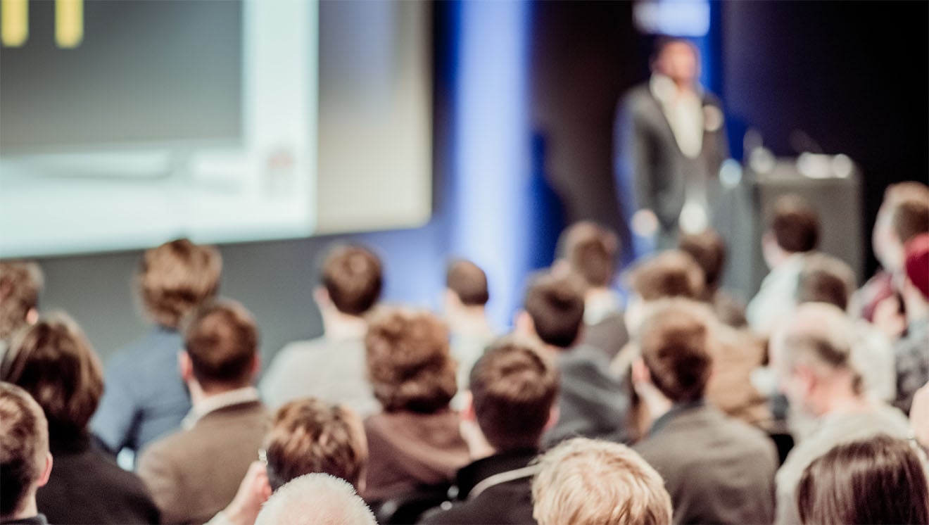 Audience watching a speaker presentation on stage at a conference.