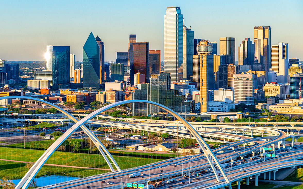 Vibrant aerial view of the Dallas skyline featuring the iconic Reunion Tower and modern skyscrapers rising above a network of sweeping highway overpasses on a bright, clear day. - Saviynt