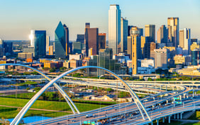 Vibrant aerial view of the Dallas skyline featuring the iconic Reunion Tower and modern skyscrapers rising above a network of sweeping highway overpasses on a bright, clear day. - Saviynt