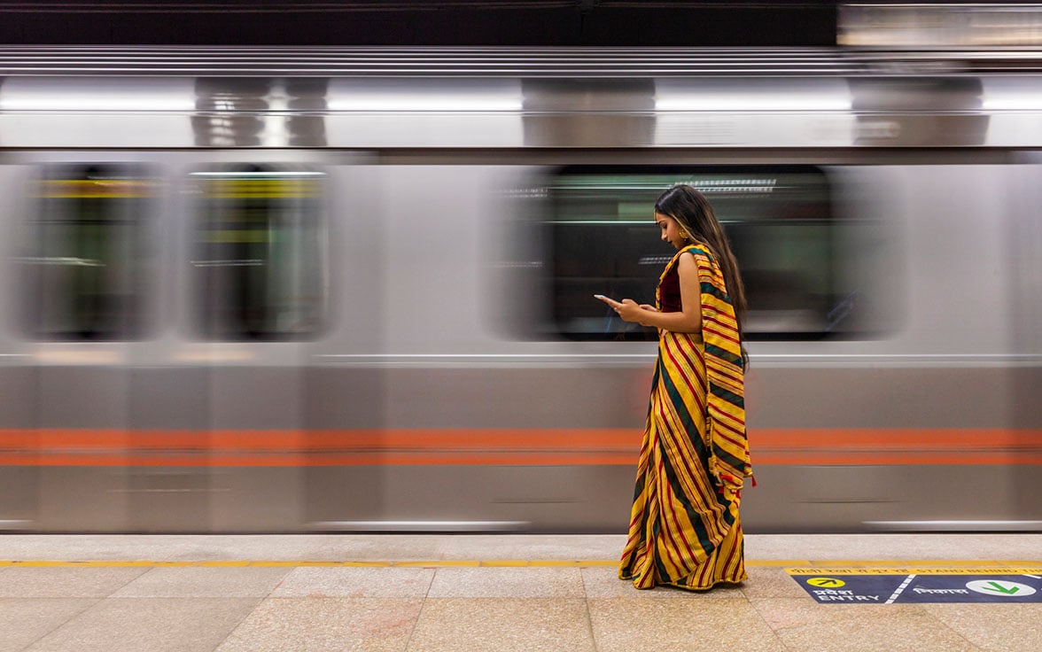 A woman wearing a vibrant red and yellow striped saree stands on a subway platform, looking at her smartphone while a blurred metro train speeds past in the background. - Saviynt