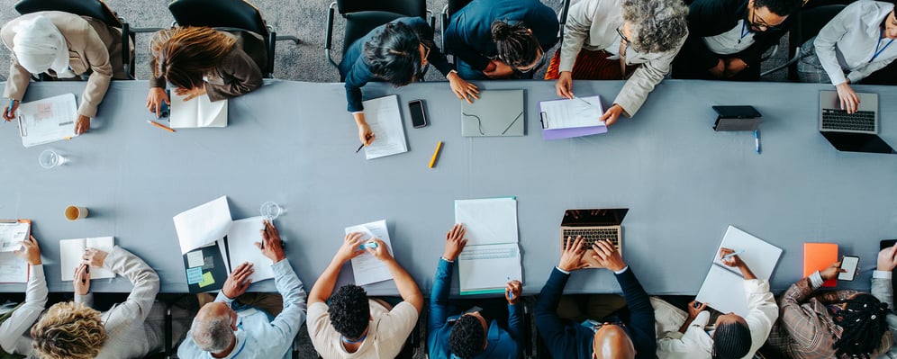 Top-down view of a diverse group of corporate professionals seated around a large conference table collaborating with laptops, notebooks, and documents during a strategic business meeting - Saviynt