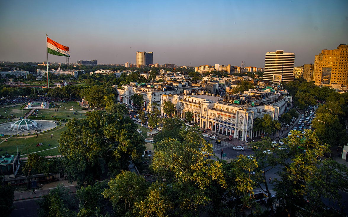 Aerial view of Connaught Place in New Delhi, India, featuring the Indian national flag in Central Park surrounded by circular architecture, lush trees, and city buildings under a clear sky.