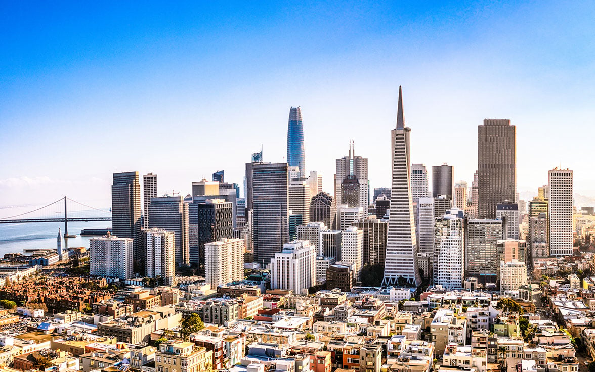 Sun-drenched aerial view of the San Francisco skyline, showcasing the iconic Transamerica Pyramid and Salesforce Tower rising above the city, with the Bay Bridge visible in the distance under a clear blue sky. - Saviynt