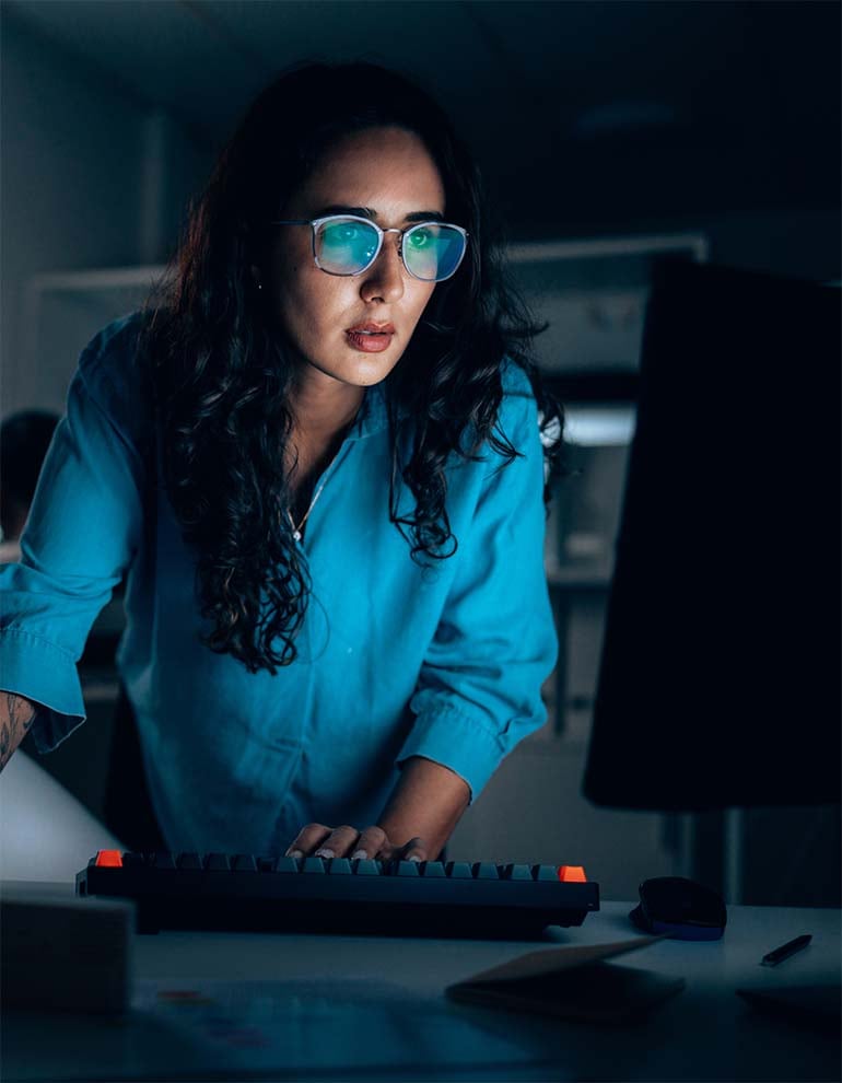 A professional woman with glasses and long dark hair working on a computer in a dimly lit room with blue ambient lighting.