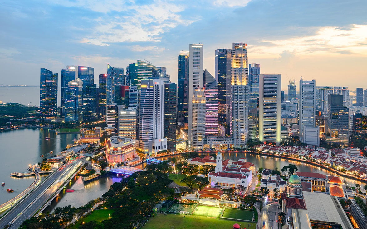 Aerial view of downtown Singapore's civic district and city skyline at dusk, featuring illuminated historic buildings, green lawns, and surrounding skyscrapers.