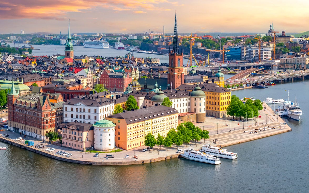 Aerial view of the Stockholm skyline featuring the historic buildings of Riddarholmen island, the iconic spire of Riddarholmen Church, and boats docked along the waterfront under a warm sunset sky. - Saviynt