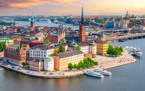 Aerial view of the Stockholm skyline featuring the historic buildings of Riddarholmen island, the iconic spire of Riddarholmen Church, and boats docked along the waterfront under a warm sunset sky. - Saviynt