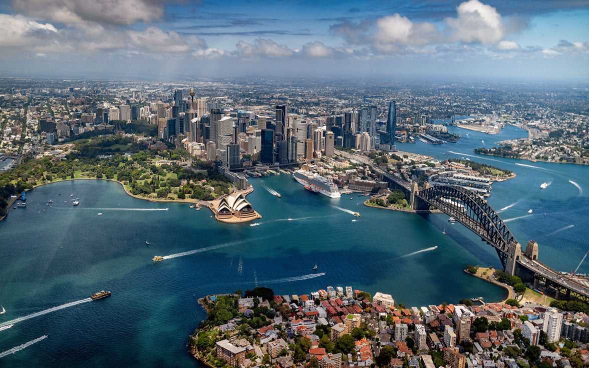 Spectacular aerial view of Sydney Harbour, featuring the iconic Sydney Opera House, Harbour Bridge, and the downtown skyscraper skyline under a partly cloudy blue sky. - Saviynt