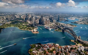 Spectacular aerial view of Sydney Harbour, featuring the iconic Sydney Opera House, Harbour Bridge, and the downtown skyscraper skyline under a partly cloudy blue sky. - Saviynt