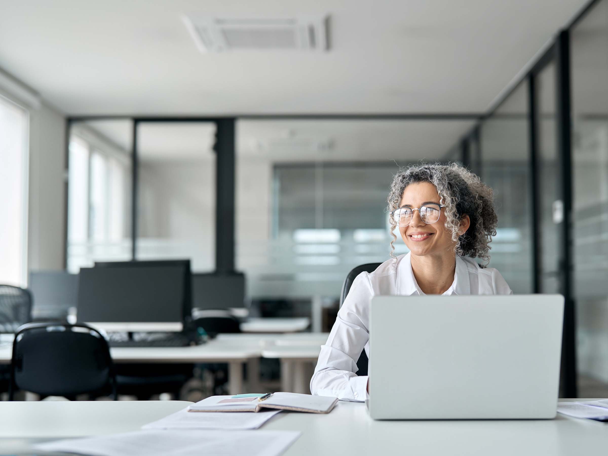 A professional woman with glasses and a white sweater smiling while working on a laptop in a modern office.